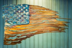 Battled Colors is metal wall art design depicting a tattered American Flag made out of metal. This particular flag has a multi-color patina which highlights the area around the cut out stars blue while the stripes are a woodgrain copper look.