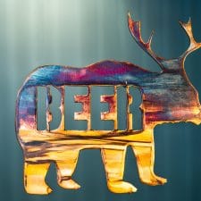 Metal art of a bear body with deer antlers and the word "beer" in the body of the bear - all cut out of metal. This piece has a multi-color patina finish with dark blues and purples up top and golden yellow towards the feet.