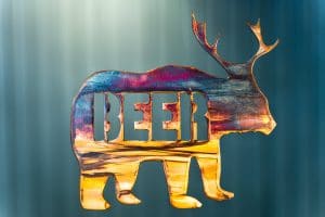 Metal art of a bear body with deer antlers and the word "beer" in the body of the bear - all cut out of metal. This piece has a multi-color patina finish with dark blues and purples up top and golden yellow towards the feet.