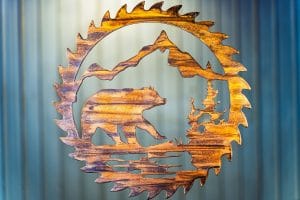 Bear Saw Blade Metal Wall Art depicts a mountain scene with a bear walking on all fours within a saw blade outline. This has a Wood Grain Copper Patina finish with some Multi-Color elements.