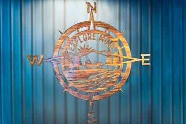 Explore More Compass metal wall decor is a large metal cutout of a compass with a class Jeep in the middle with the phase Explore More. The metal is finished with a wood grain copper patina.