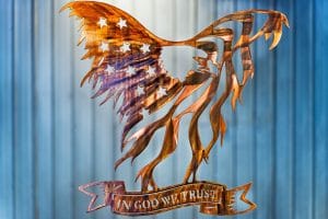 Metal wall art of American Bald Eagle with expanded wings depicting American Flag and holding In God We Trust banner in talons all cut out of metal with a Wood Grain Copper Patina.