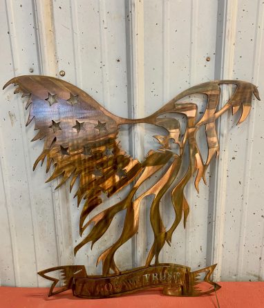 Metal wall art of American Bald Eagle with expanded wings depicting American Flag and holding In God We Trust banner in talons all cut out of metal with a Wood Grain Copper Patina. This picture shows Freddie Freedom at the shop.