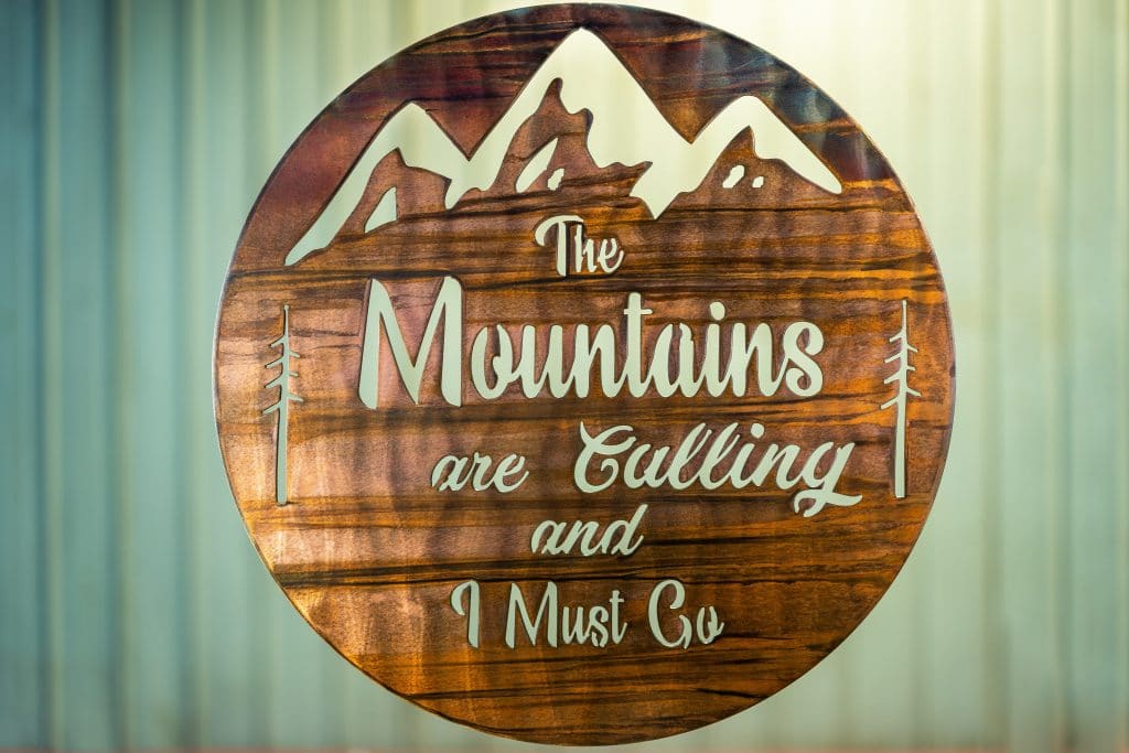 The Mountains are calling and I must go metal wall art is a circular piece of metal with the phrase cut into the metal with mountains in the background.
