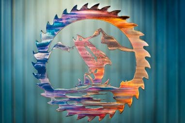 Metal wall art cut out of a saw blade shape depicting at trout jumping out of the water with a mountain in the background inside the saw blade. This has a Multi-Color Patina finish with deep blues and purples along side wood grain copper effects.