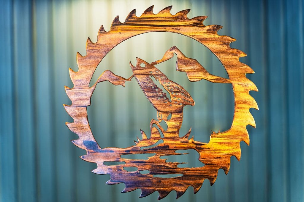 Metal wall art cut out of a saw blade shape depicting at trout jumping out of the water with a mountain in the background inside the saw blade. This has a Wood Grain Copper Patina finish with some Multi-Color elements