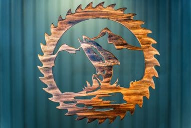 Metal wall art cut out of a saw blade shape depicting at trout jumping out of the water with a mountain in the background inside the saw blade. This has a Wood Grain Copper finish.