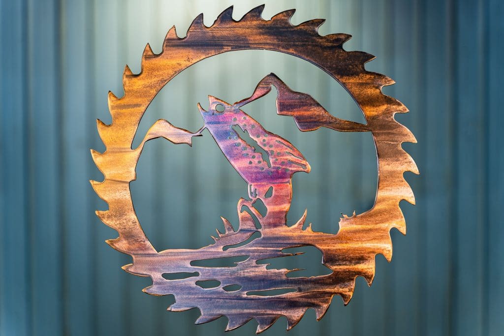 Metal wall art cut out of a saw blade shape depicting at trout jumping out of the water with a mountain in the background inside the saw blade. This has a Multi-Color Patina finish with deep blues and purples along side wood grain copper effects.