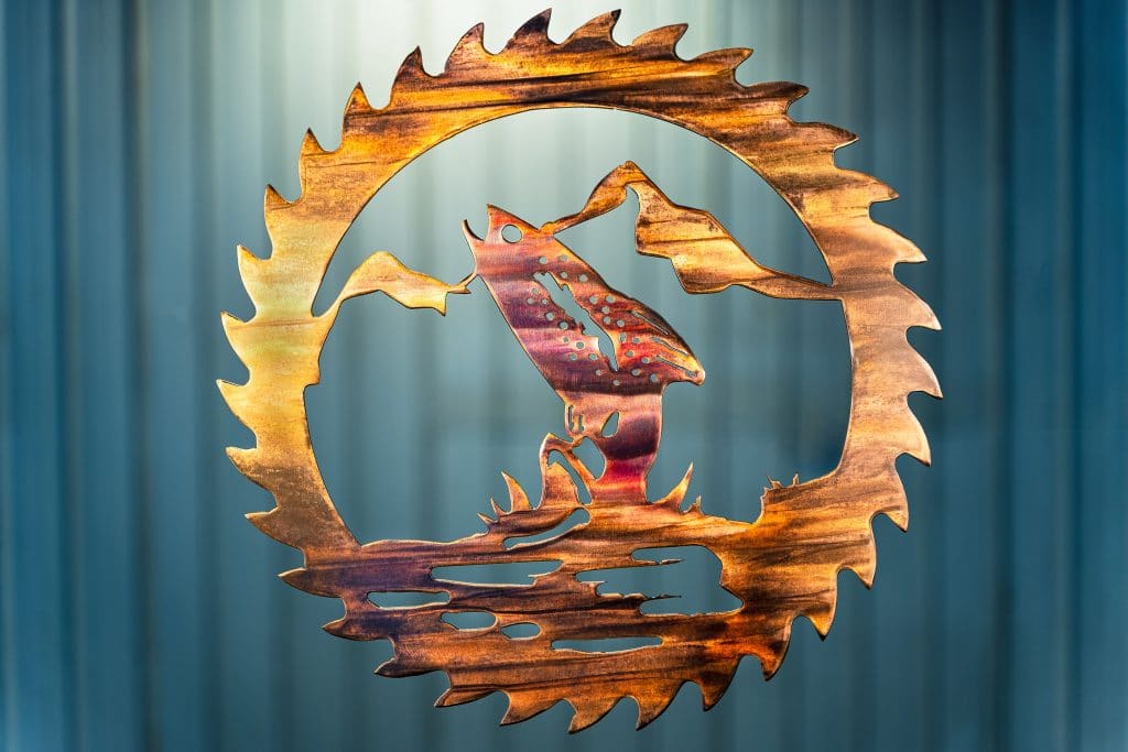 Metal wall art cut out of a saw blade shape depicting at trout jumping out of the water with a mountain in the background inside the saw blade. This has a wood grain copper patina with some multi-color effects.