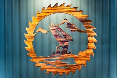 Metal wall art cut out of a saw blade shape depicting at trout jumping out of the water with a mountain in the background inside the saw blade. This has a Wood Grain Copper finish.