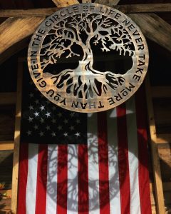 Circular metal wall art with a tree in the middle and the phrase In The Circle of Life Never Give More Than You Take. This picture shows the metal wall art projecting a shadow of the design onto an American Flag