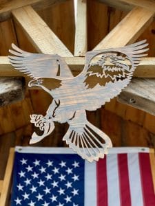 Metal wall art of American Bald Eagle with expanded wings and fish in talons all cut out of metal with a Wood Grain Copper Patina. There is also a portrait of a bald eagle in the right week cut out of metal. This picture shows the metal wall decor hanging on a cabin with an American flag in the background