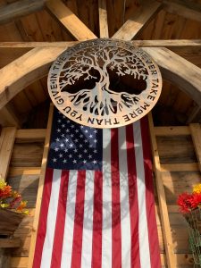 Circular metal wall art with a tree in the middle and the phrase In The Circle of Life Never Give More Than You Take. This picture shows the metal wall art projecting a shadow of the design onto an American Flag