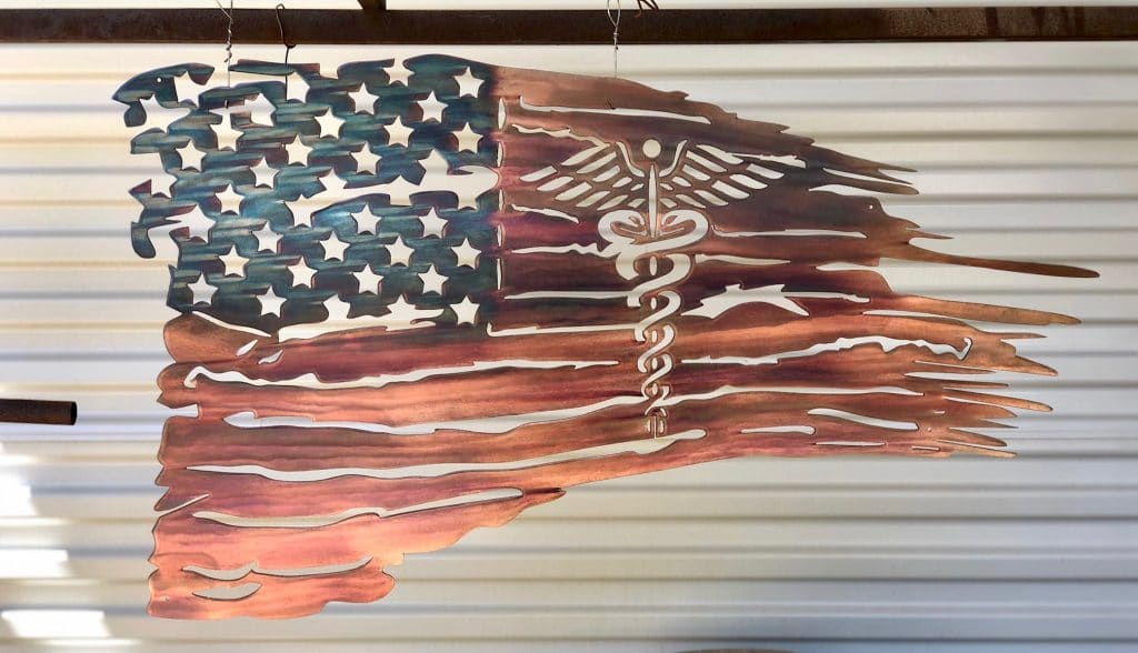 The New Battlefield Tattered Flag metal art depicts a ragged version of the American flag with the medical caduceus symbol in the flag