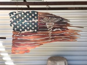 The New Battlefield Tattered Flag metal art depicts a ragged version of the American flag with the medical caduceus symbol in the flag